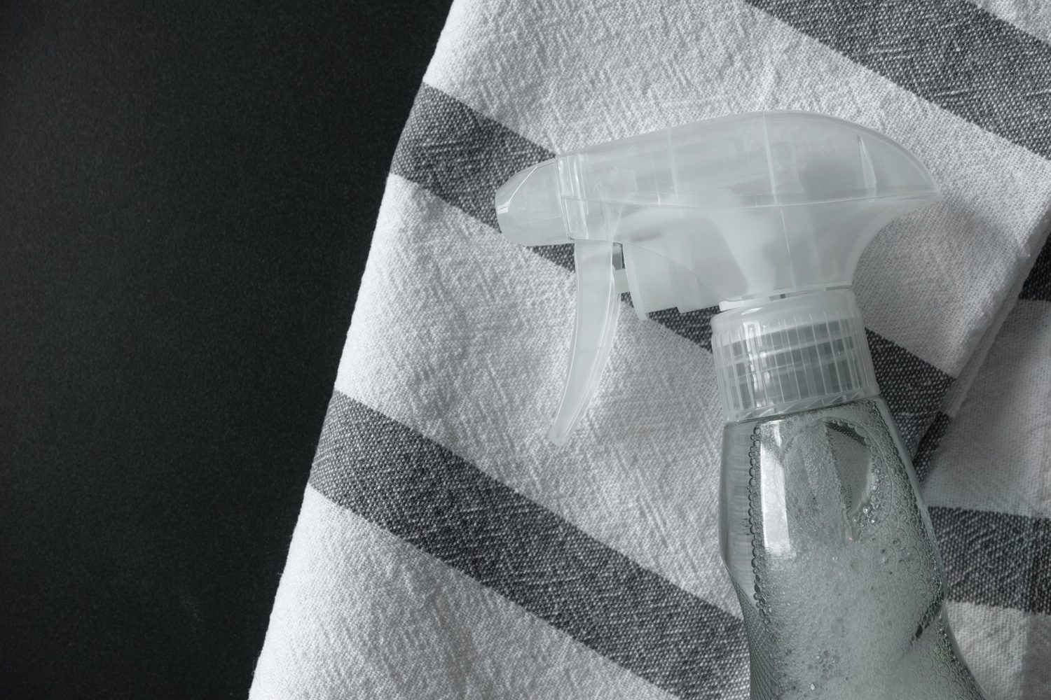 Una botella pulverizadora transparente con producto de limpieza descansa sobre un paño de rayas grises y blancas.