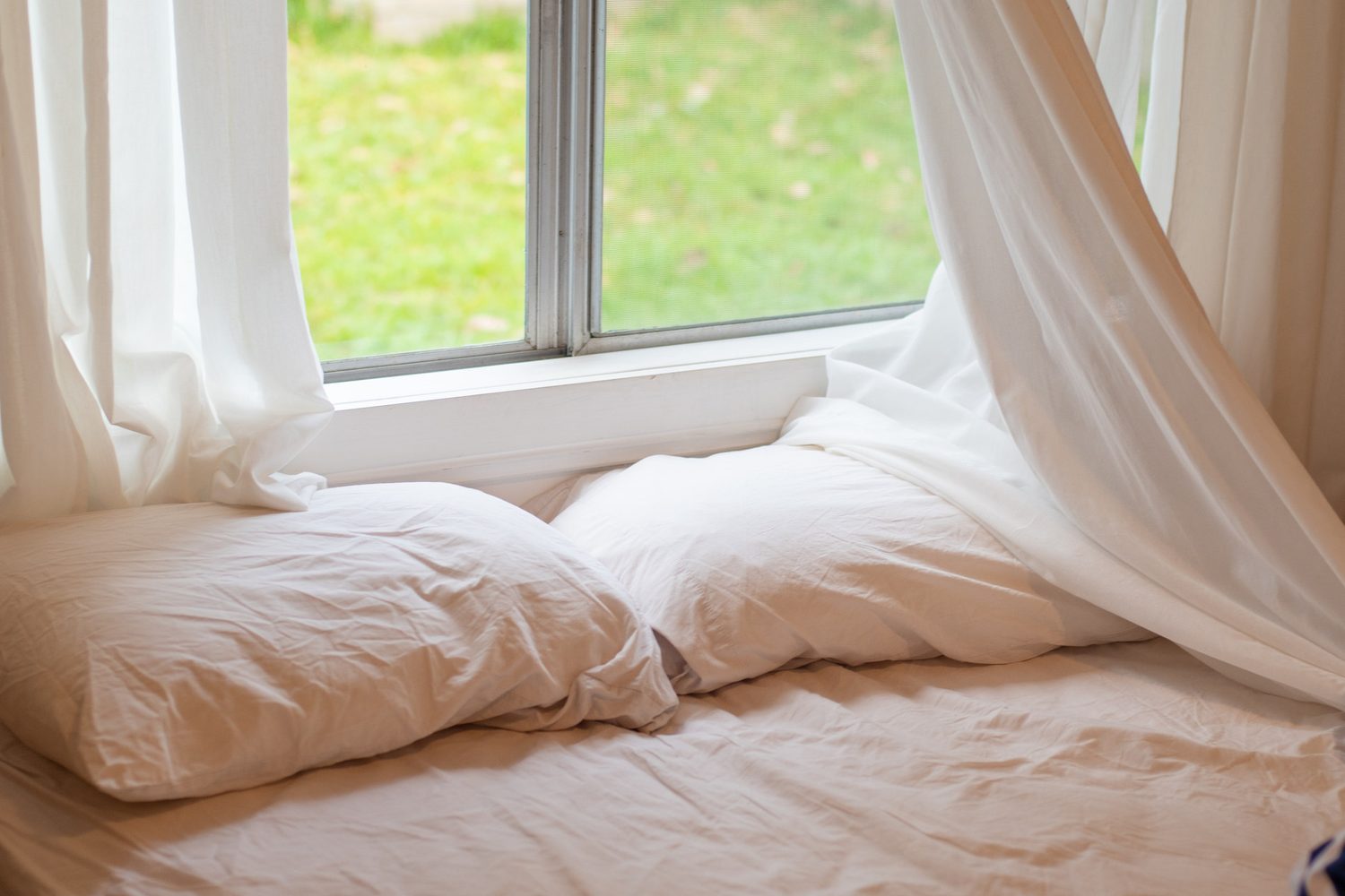 Cama con sábanas y almohadas blancas junto a una ventana iluminada por luz natural