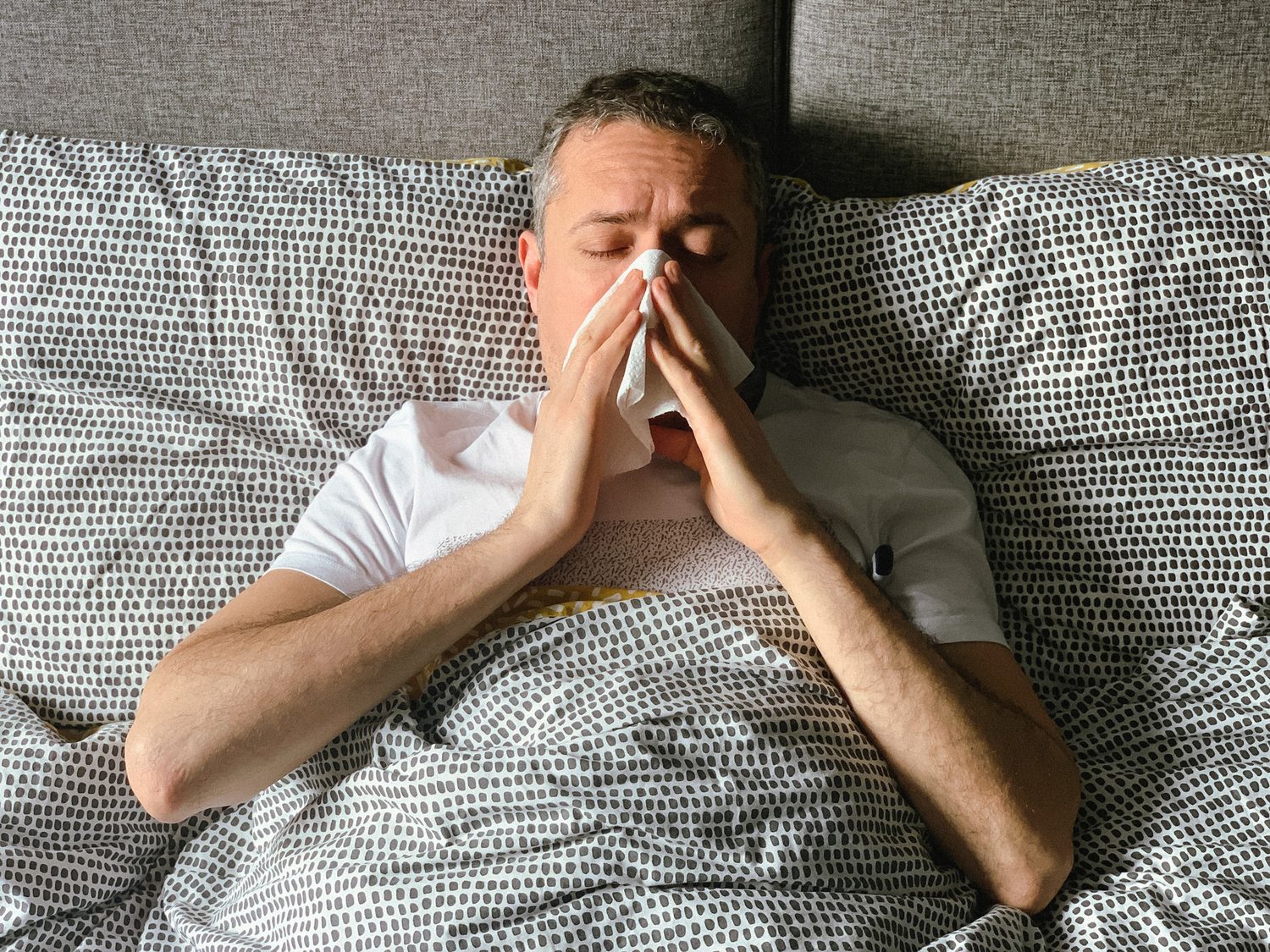 Hombre tumbado en la cama sonándose la nariz con un pañuelo entre almohadas estampadas