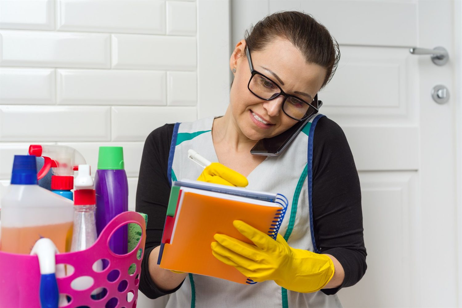 Limpiadora con guantes amarillos anota tareas en una libreta mientras habla por el móvil junto a sus productos de limpieza