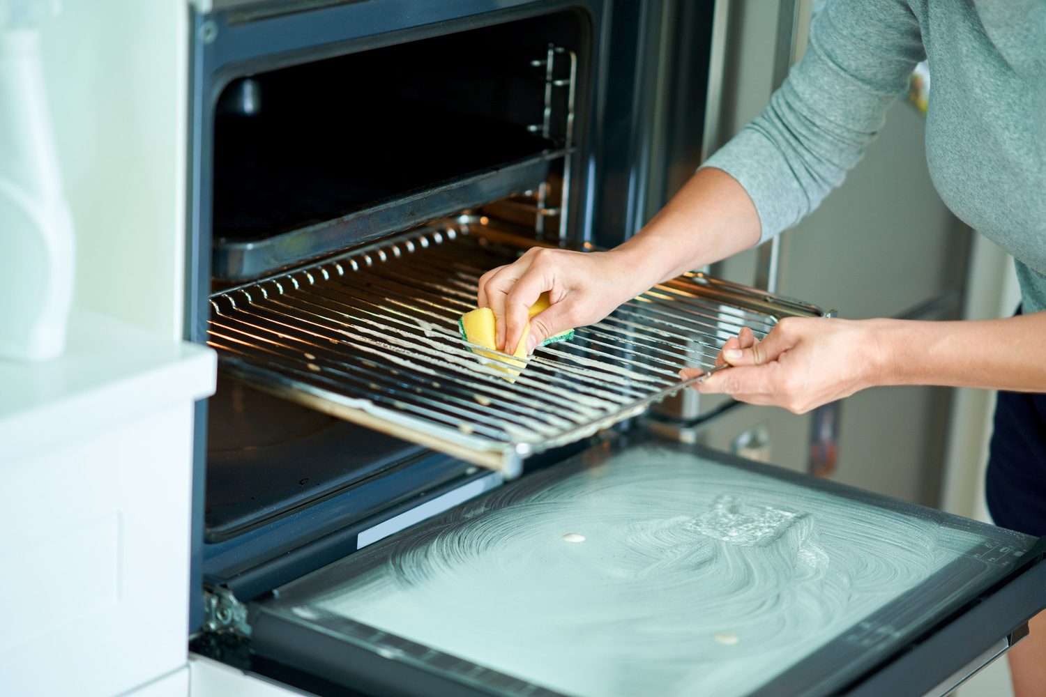 Persona limpiando la rejilla de un horno abierto con una esponja amarilla