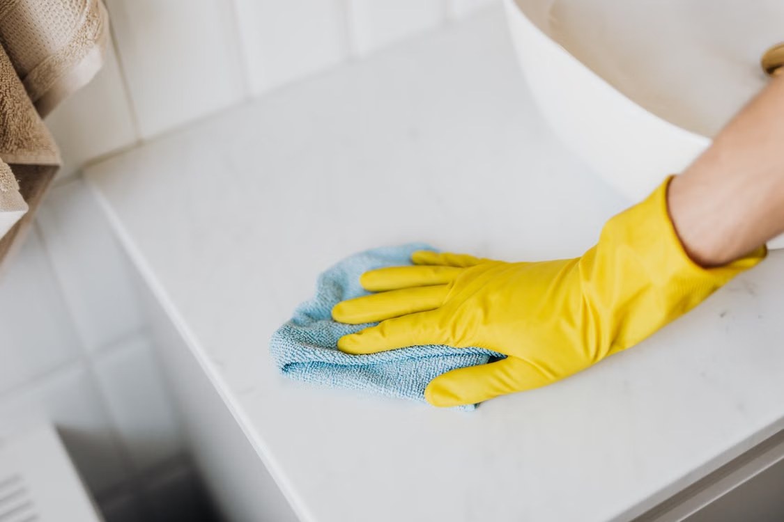 Mano con guante amarillo limpia una encimera de mármol blanco con un paño de microfibra azul.