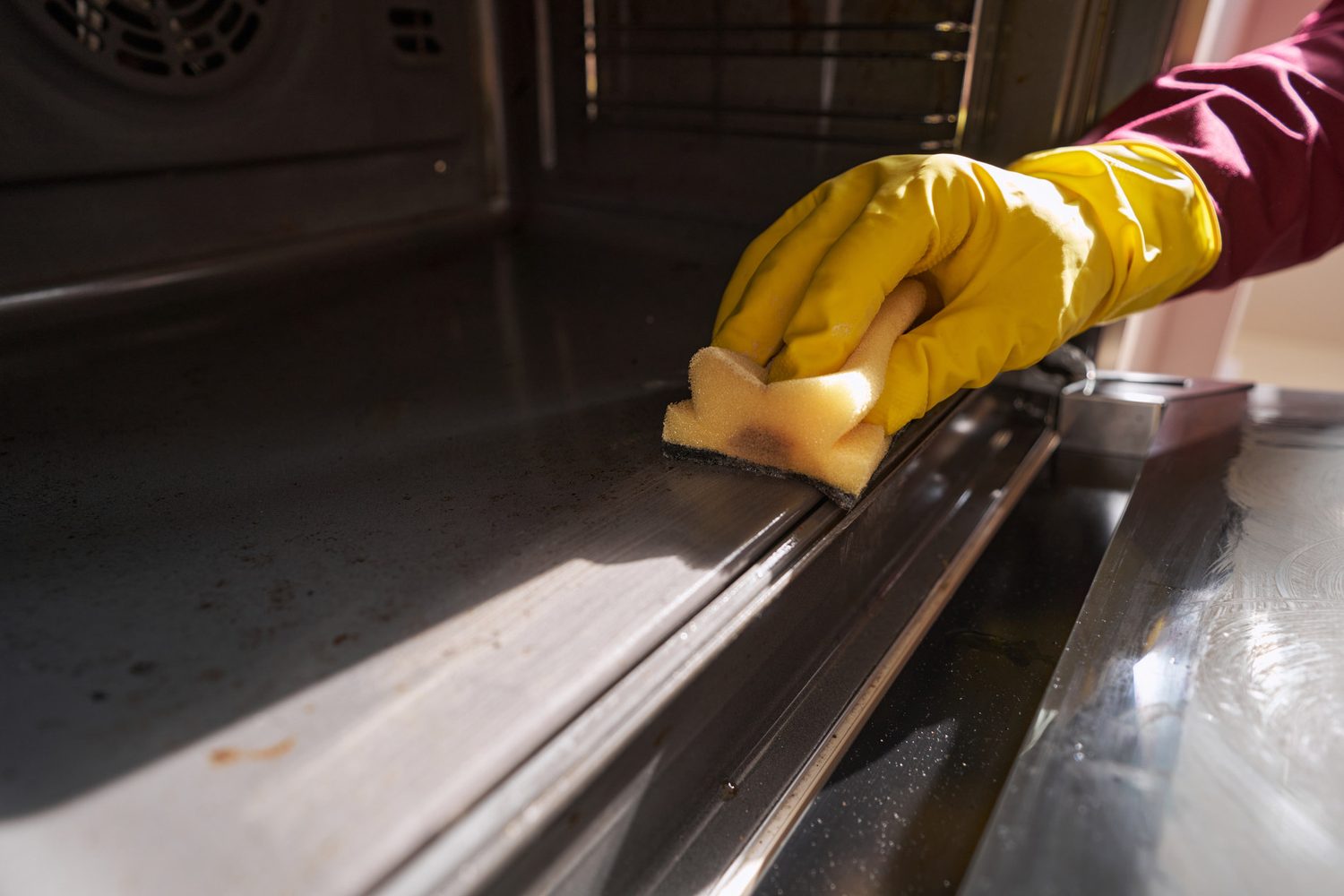 Mano con guante amarillo que limpia el interior de un horno abierto con una esponja