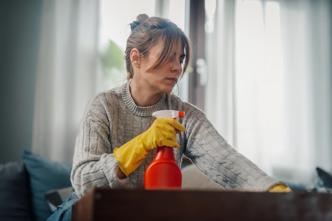 Una mujer con guantes amarillos limpia una superficie de casa mientras sostiene un pulverizador rojo
