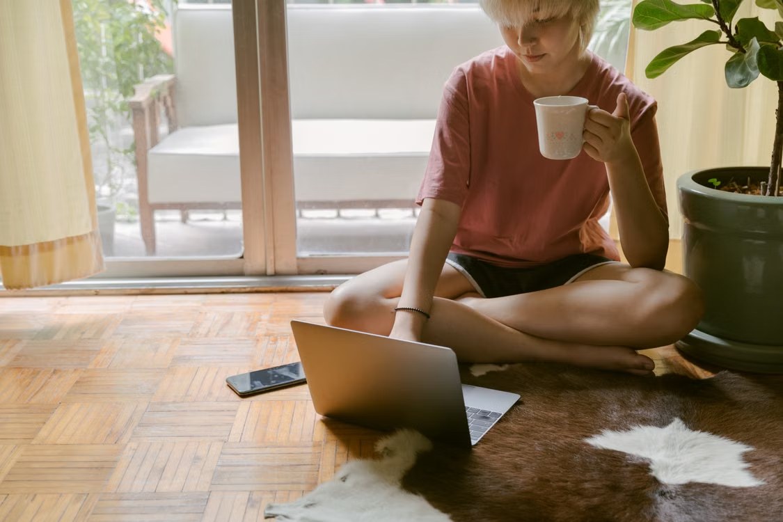 Una mujer sentada en el suelo del salón consulta su portátil mientras sostiene una taza
