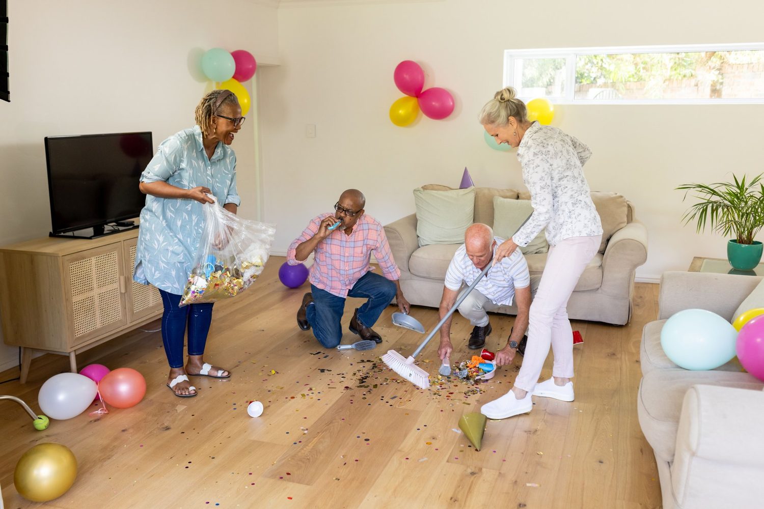 Varias personas recogen confeti y restos de una fiesta en un salón decorado con globos.
